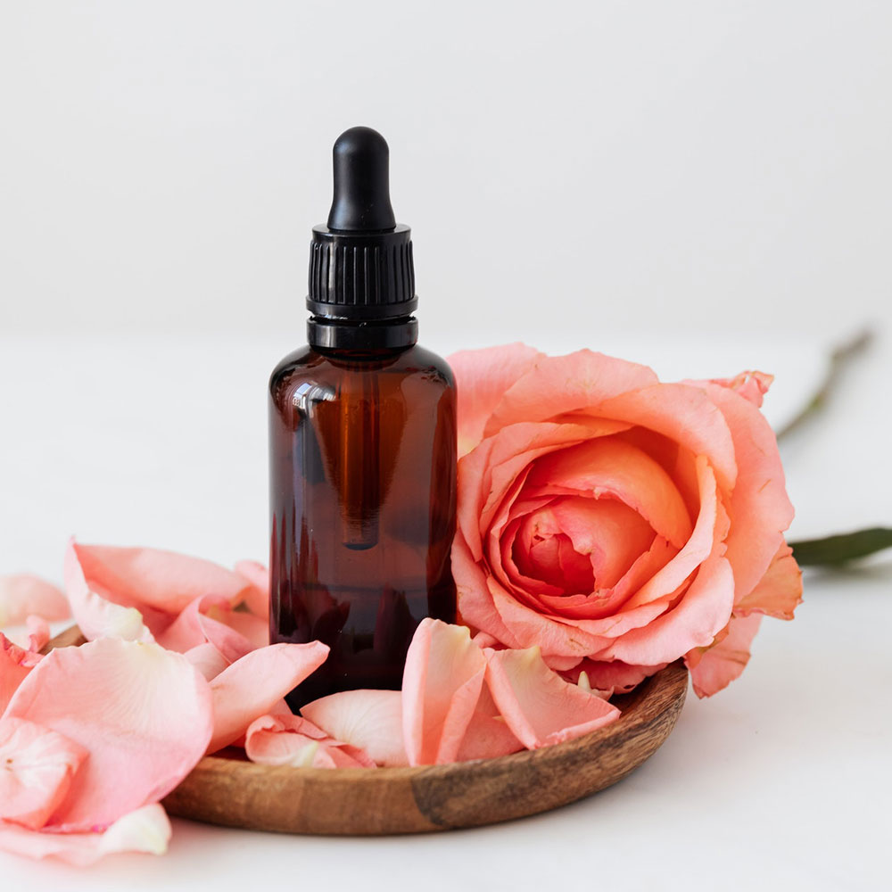 An essential oil in a dropper bottle over the wooden coaster with pink roses and petals
